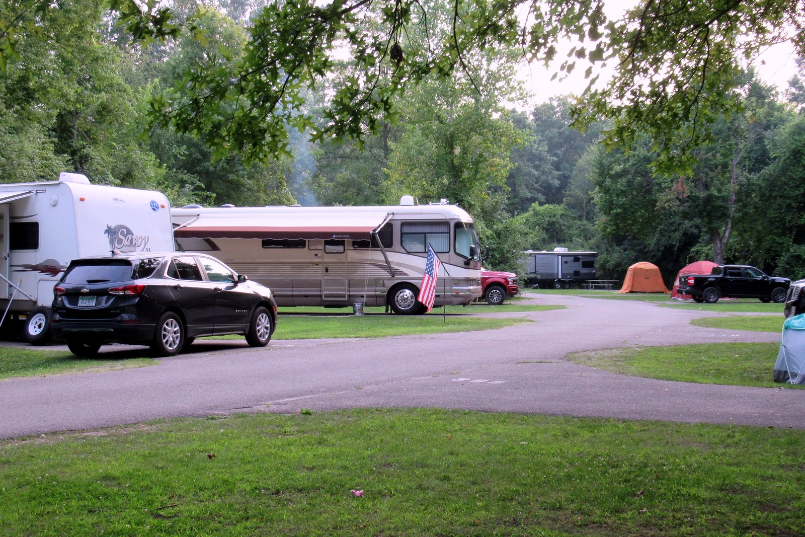 Motor homes, trailers, cars and tents fill shaded campsites along a paved loop road in a wooded campground.