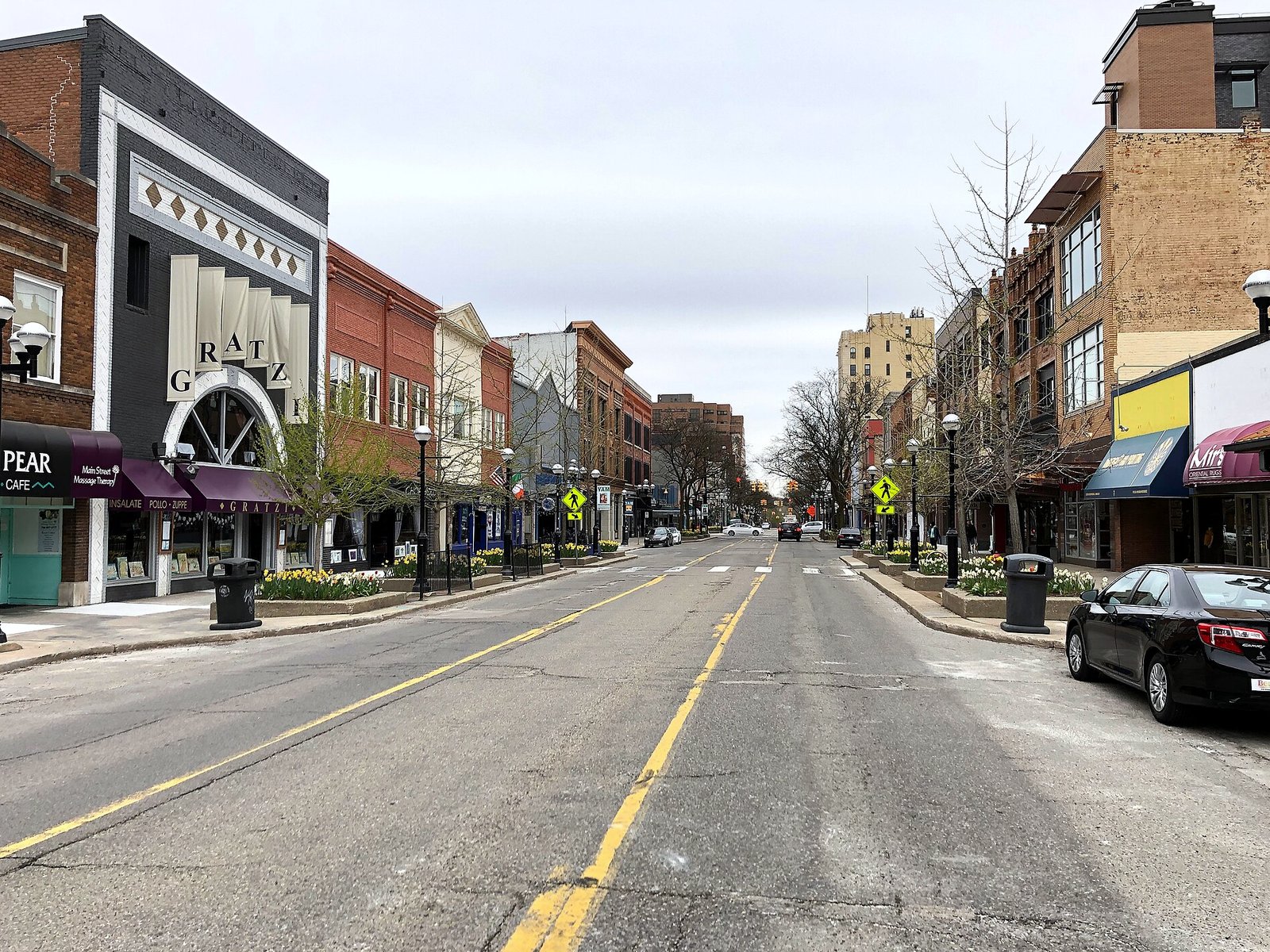A wide downtown street lined with two- and three-story brick storefronts, an arched marquee reading Gratz on the left, street lamps and planters along both sidewalks under a cloudy sky.