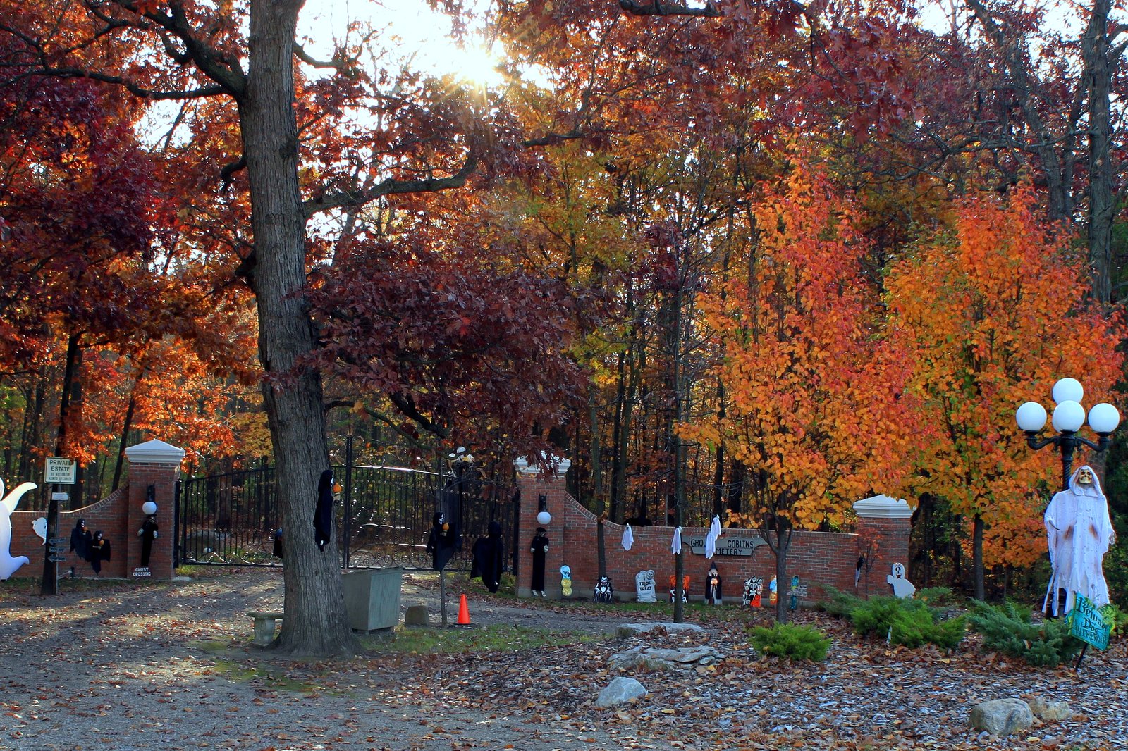 A yard decorated for Halloween with white ghost figures and small decorations along a brick-and-iron fence, under trees in bright red and orange fall color.