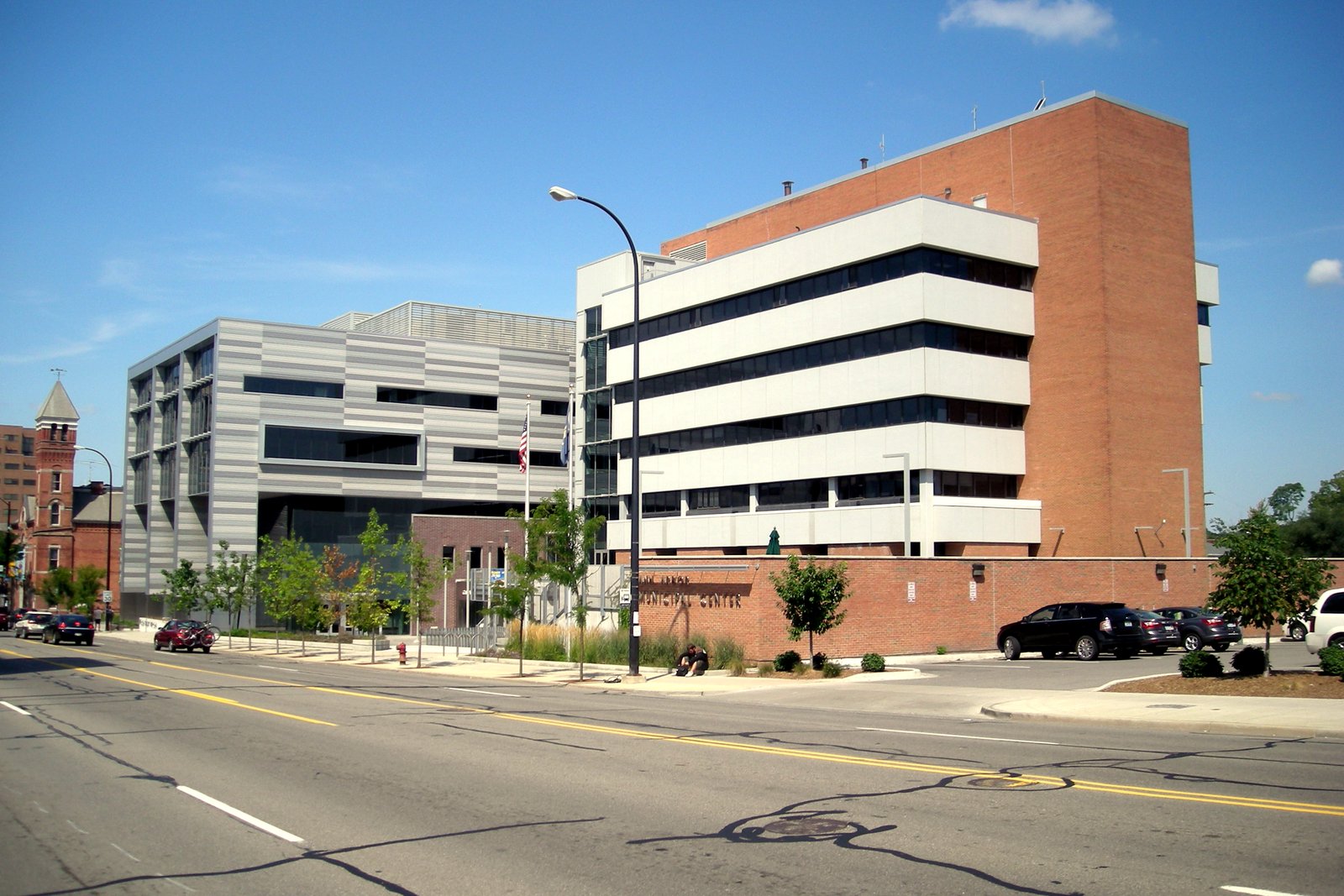 A modern multistory city hall building with horizontal bands of windows and a brick tower beside a wide downtown street under blue sky.