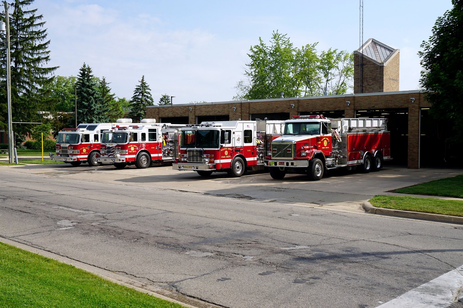 Four red fire engines parked on the apron in front of the open bays of a brick fire station in Saline, with evergreen trees and a squared brick tower.