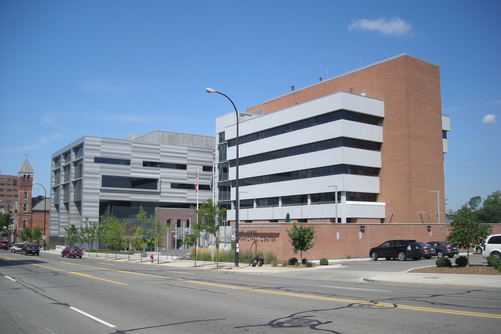 A modern gray building and a brick and white-paneled office tower behind a sign reading Municipal Center, along a patched four-lane street with parked cars.