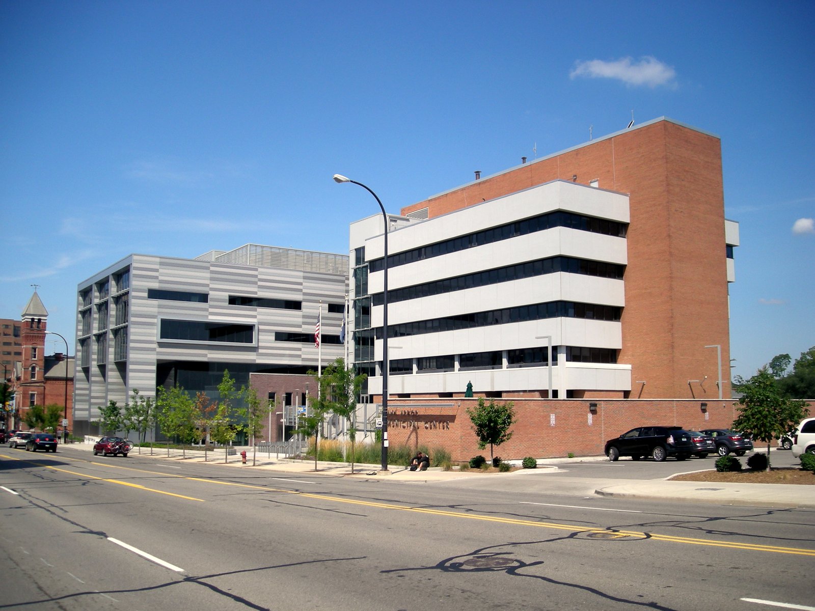 A modern municipal building with white horizontal bands and a brick tower beside a wide street under a blue sky.