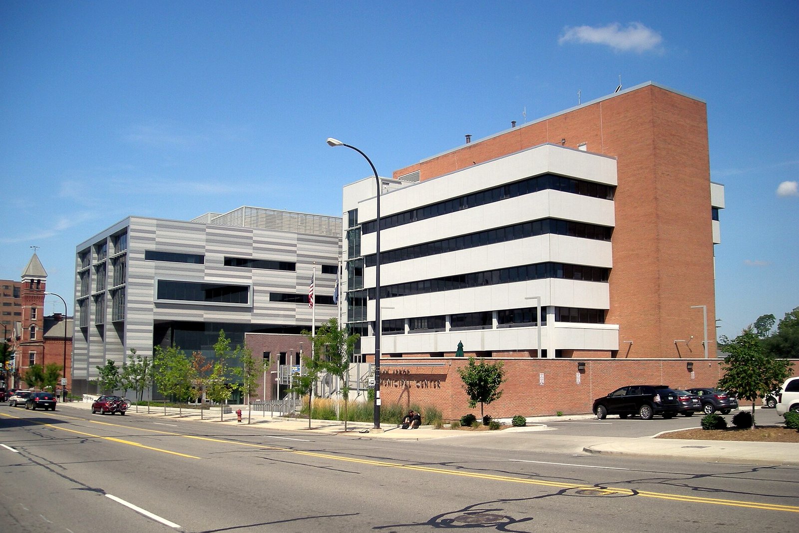 A multi-story brick and glass city hall building with a plaza and trees in front under a blue sky.