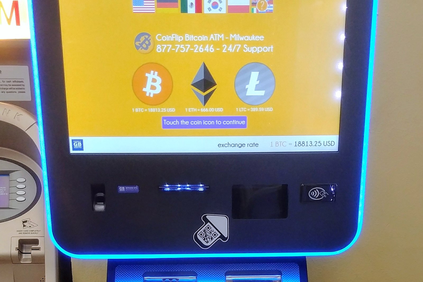 A cryptocurrency kiosk with a large yellow touch screen showing Bitcoin, Ethereum and Litecoin symbols, a lit blue frame and a cash slot below.