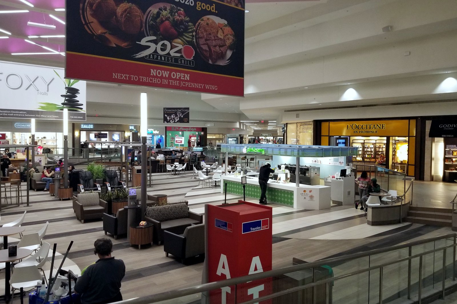 The inside of a shopping mall with a food court seating area, a lower level of shops and a large food advertisement hanging overhead.