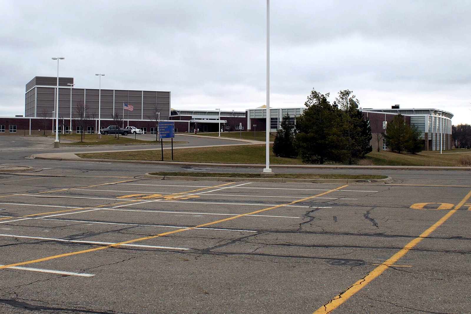 The long, low brick and glass buildings of Chelsea High School across a wide, empty parking lot under a gray sky, with an American flag flying near the entrance.