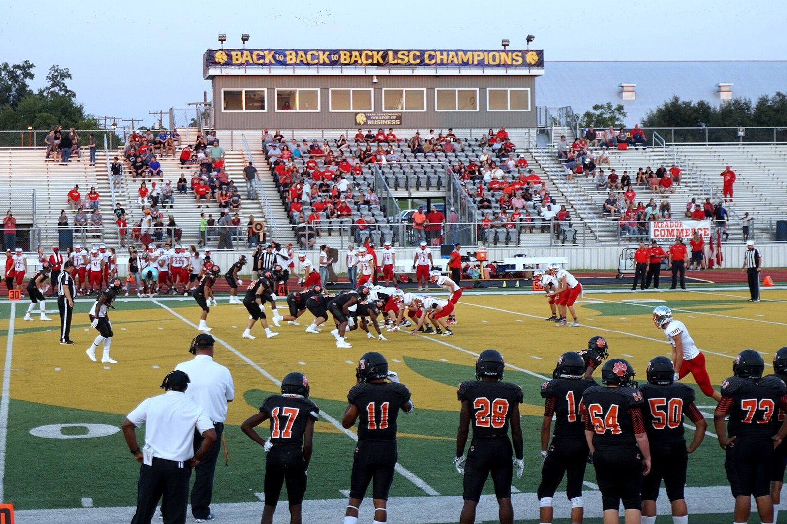 High school football players in dark jerseys line up on offense on a lighted field as defenders wait across the line of scrimmage.