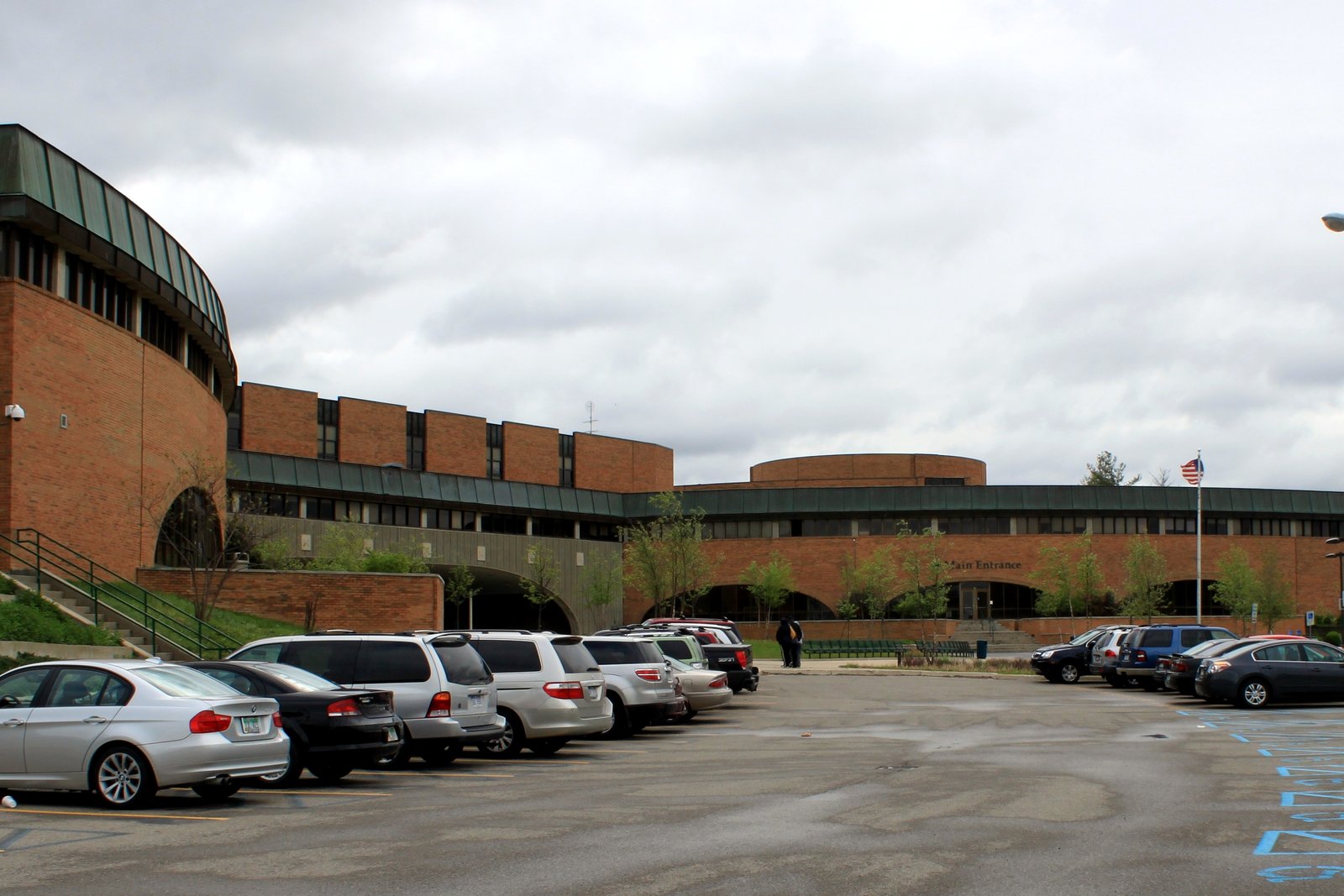 The brick exterior of Huron High School in Ann Arbor, with a curved, green-roofed wing at left, a flagpole and parked cars in front, under an overcast sky.