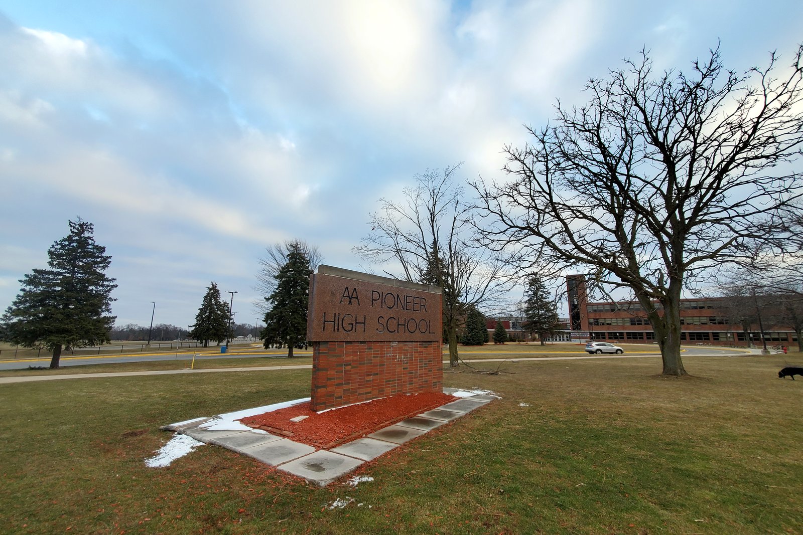 A brick school sign on a grassy lawn in front of a long, low school building, with bare trees and evergreens under a cloudy sky.