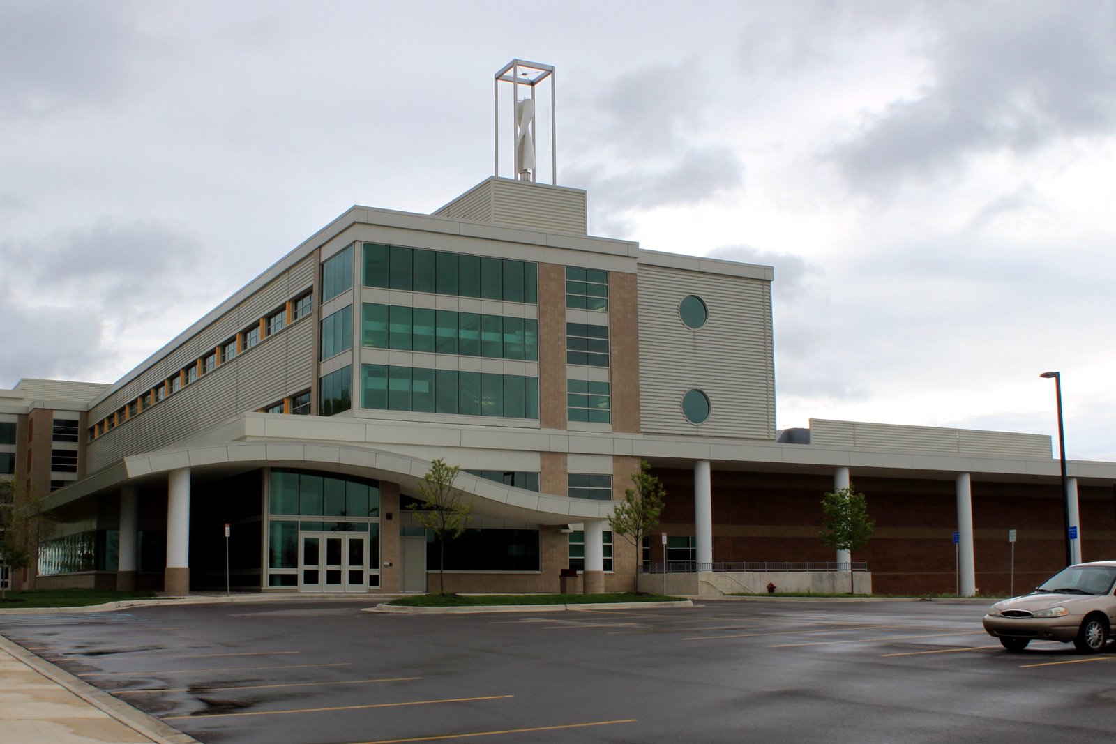A wide two-story high school building with a flat roof, parking lot and landscaping under a bright sky.