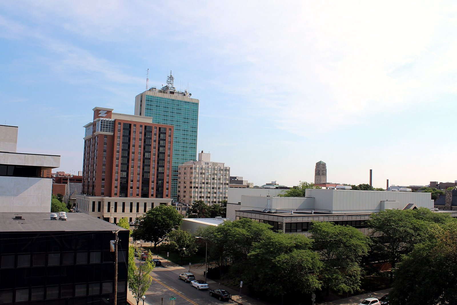 Downtown Ann Arbor seen from a parking structure on a sunny summer day, with a tall blue-green glass office tower, a red brick high-rise, a pale apartment building, low flat-roofed buildings, a tree-lined street with parked cars and a stone tower in the distance.