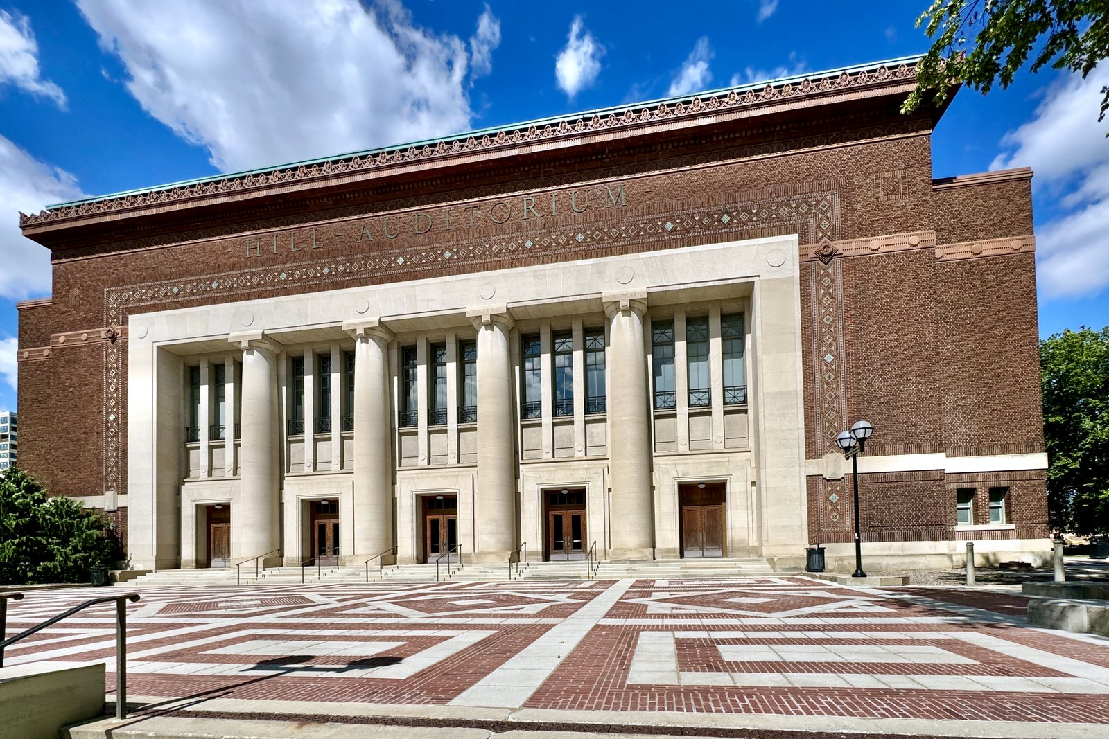 A large brick concert hall with a row of tall stone columns and the words Hill Auditorium carved above the entrance, behind a patterned brick plaza under a blue sky.