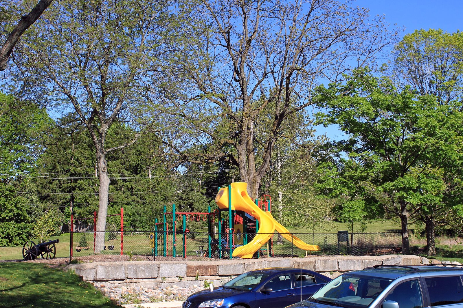Children’s playground equipment and a wood-chip play surface sit among trees in a city park on a clear day.