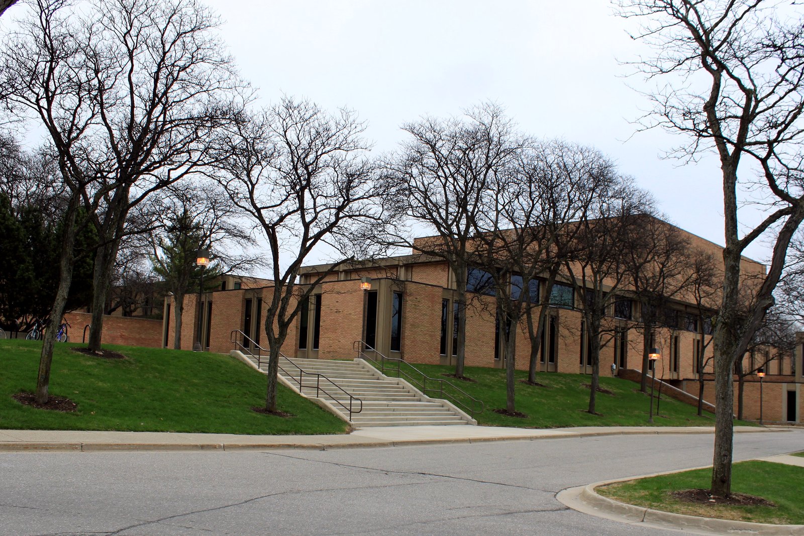 A long tan brick building with rows of windows on a grassy rise behind bare trees, with steps up from a curving road under a cloudy sky.