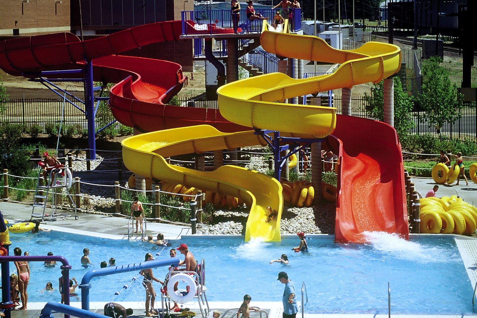 Yellow and red water slides twist down into an outdoor pool where children play on a sunny day.