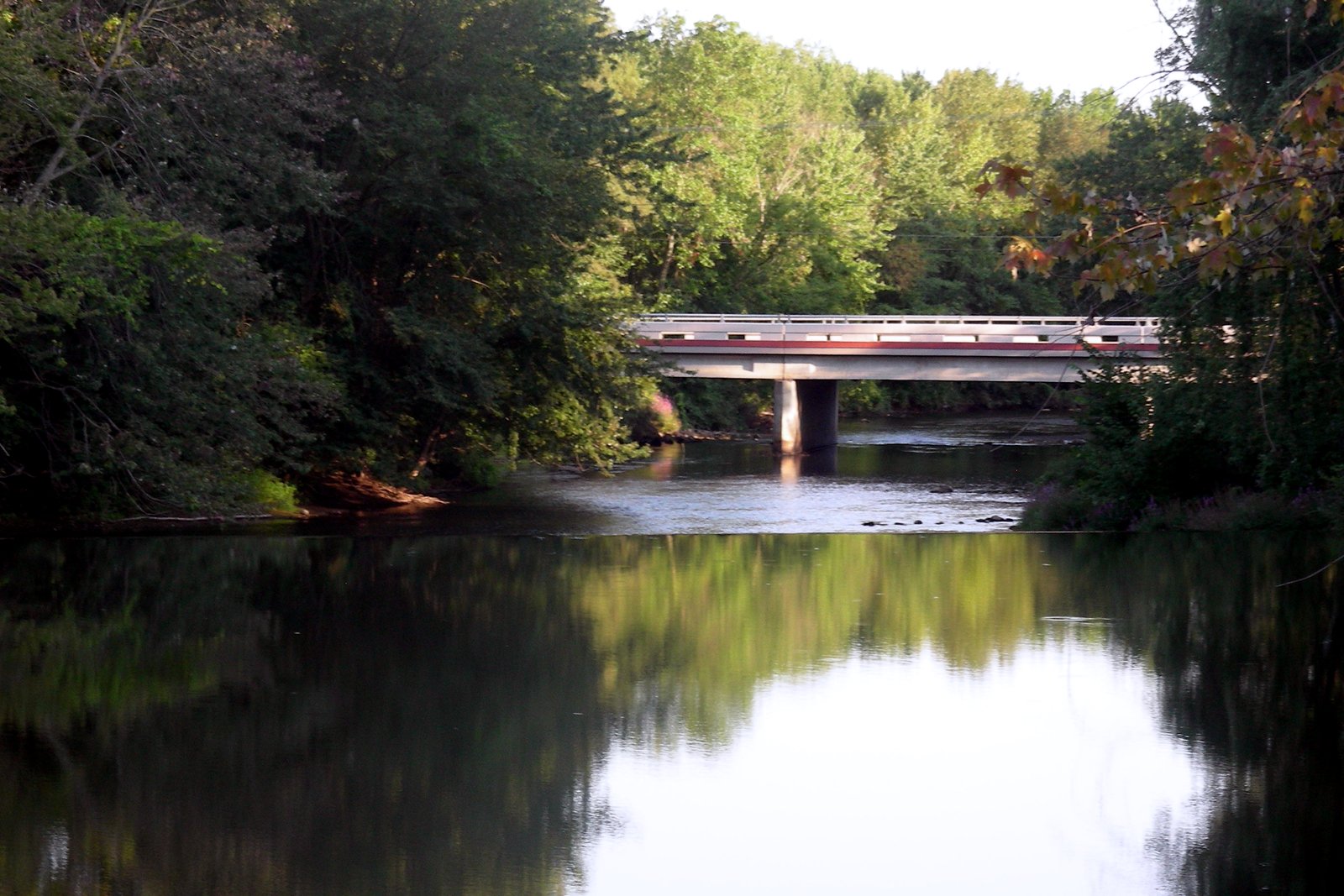 A concrete road bridge crosses a calm, tree-lined river, with the trees reflected in the water.
