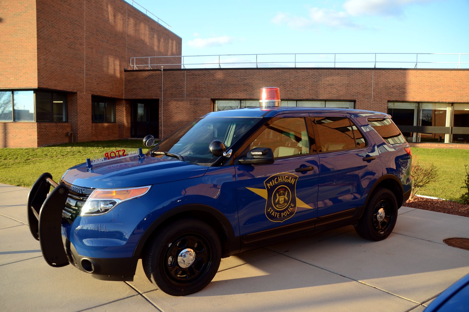 A dark blue Michigan State Police Ford Explorer parked at a curb in front of a brick building, with its push bar and light bar visible.