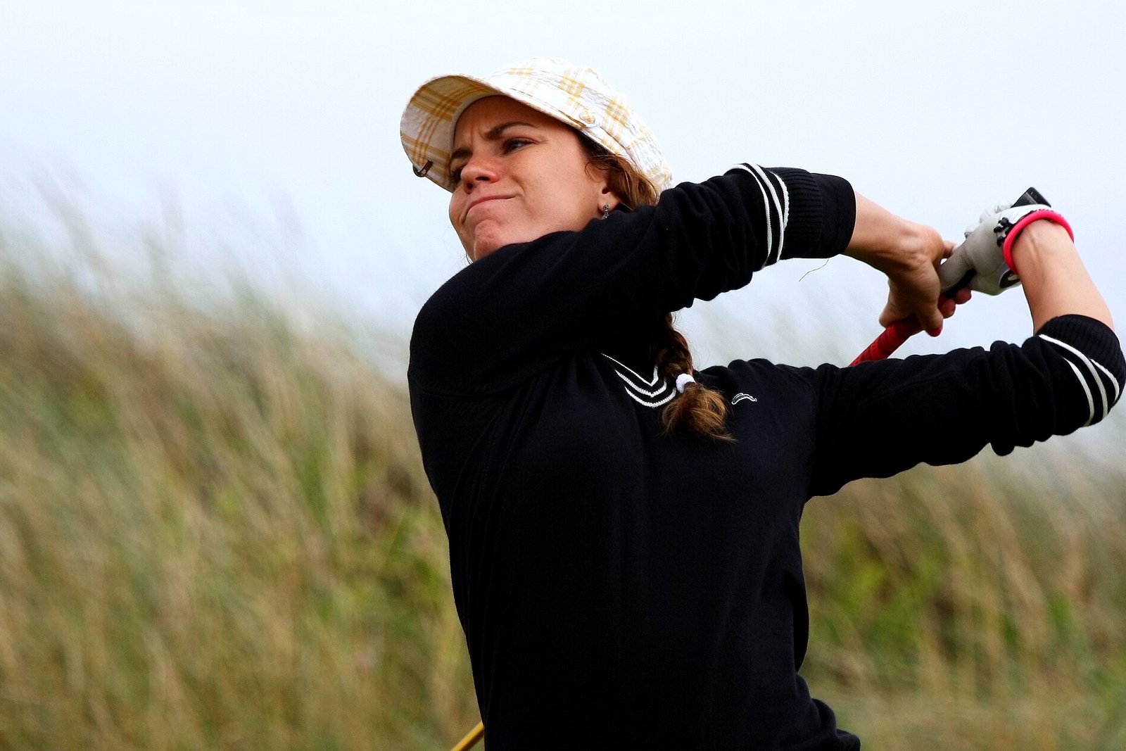 A golfer in a plaid cap and black sweater holds her follow-through and watches a tee shot, with tall dune grass behind her.
