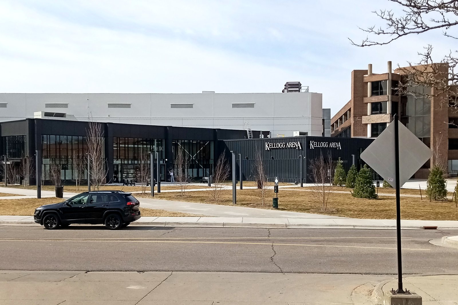 A large modern arena building with glass and metal cladding sits beside a parking lot and trees under a blue sky with scattered clouds.