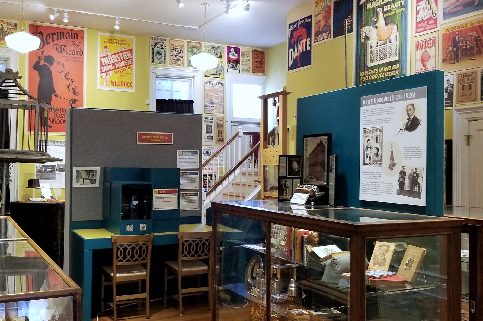A museum room crowded with glass display cases, vintage magic posters on yellow walls, a birdcage prop and an exhibit panel about Harry Houdini.