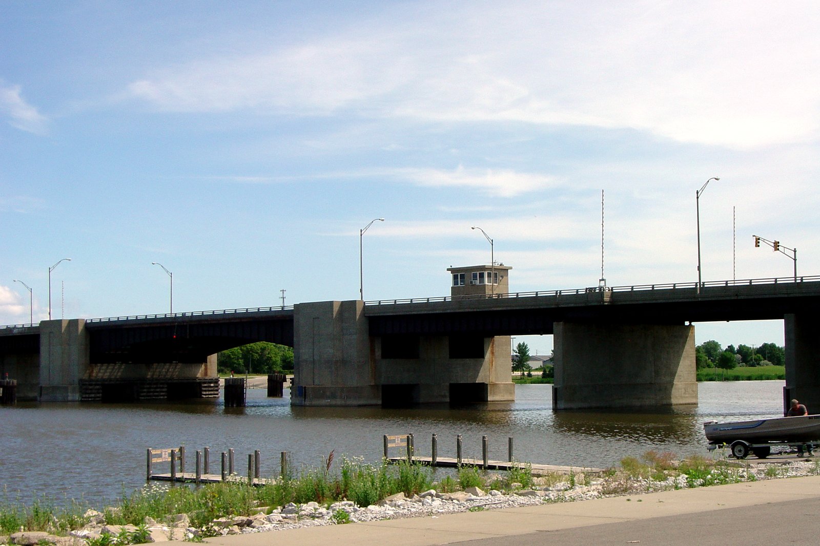 A concrete drawbridge with a small operator’s house crosses a calm river under a blue sky, with a grassy shore and boat docks in the foreground.