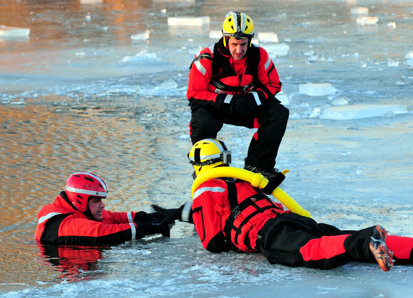 Two Coast Guard crew members in red survival suits on broken ice, one reaching for a third crew member lying on the ice.
