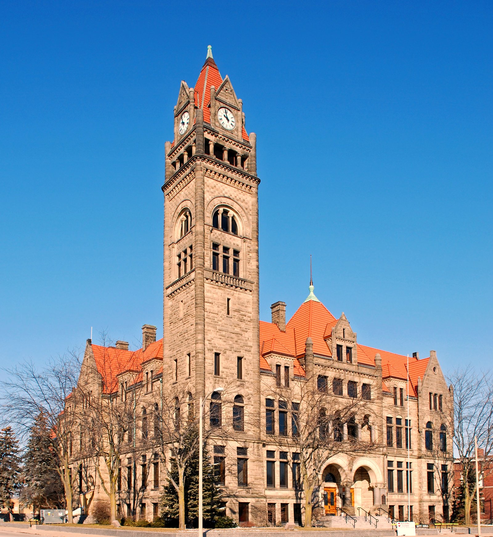 A tall stone clock tower rises beside a long Romanesque stone building with orange tile roofs under a clear blue sky, with bare trees in front.