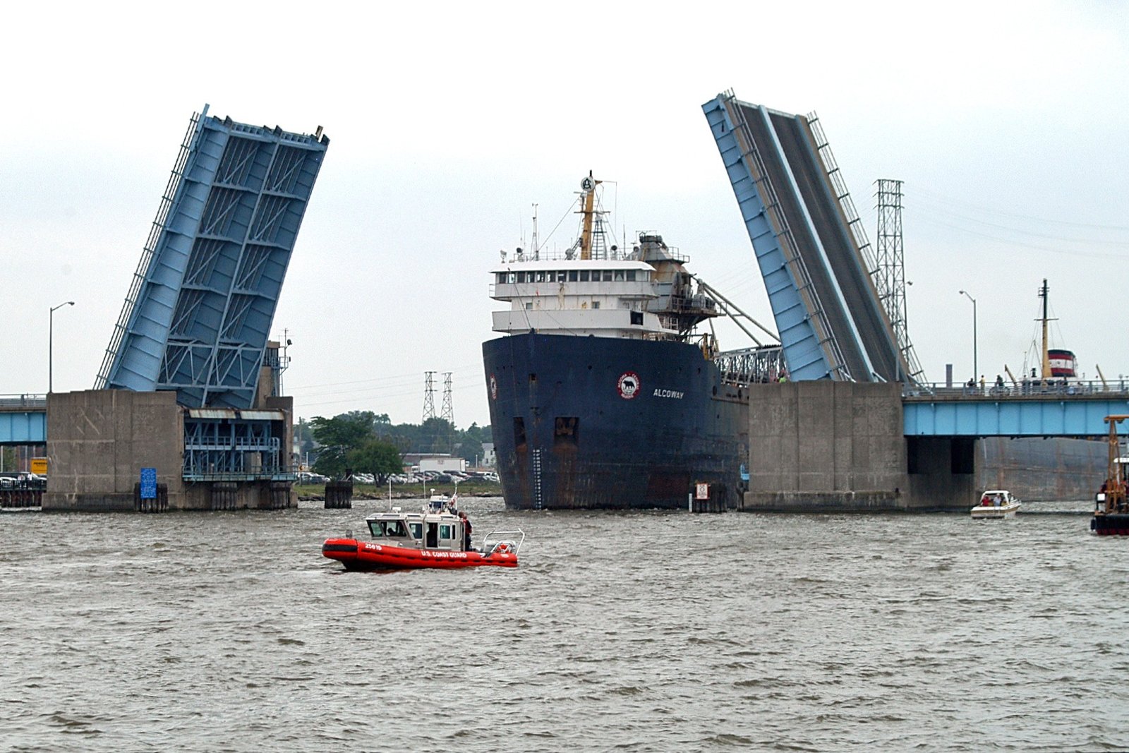 The two raised blue leaves of the Veterans Memorial Bridge drawbridge in Bay City, with the dark-hulled freighter Algoway passing through on the Saginaw River and a small orange U.S. Coast Guard boat in the foreground.