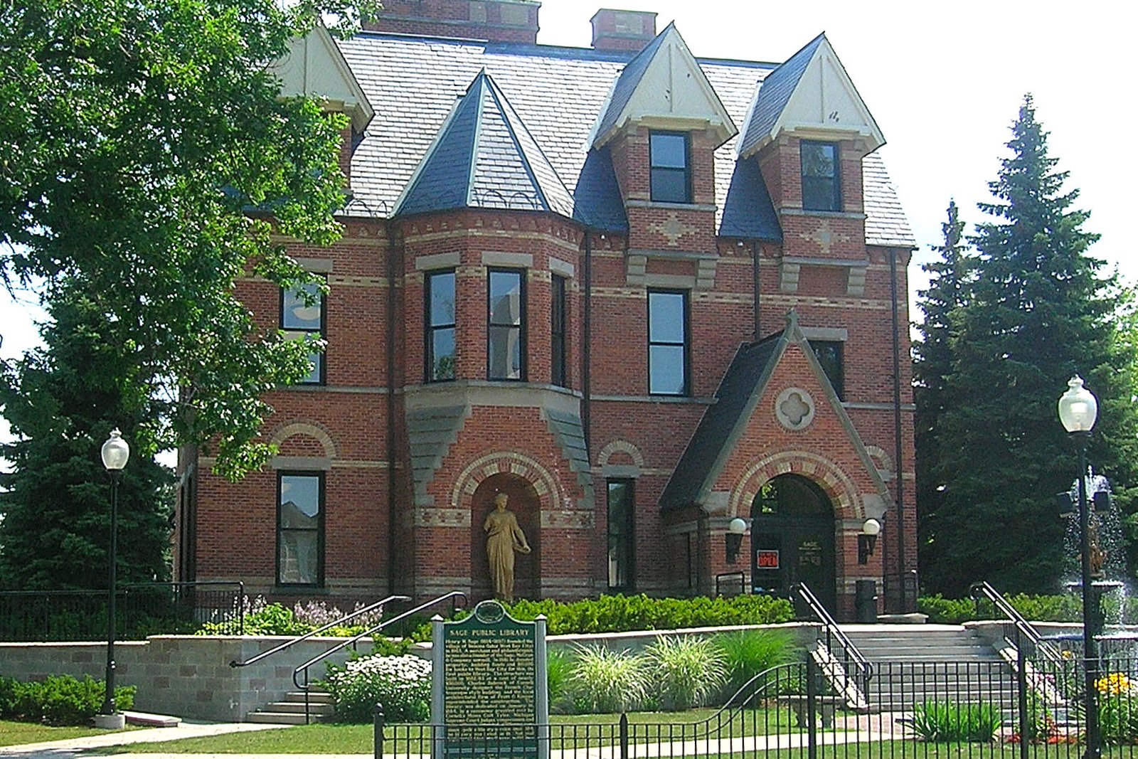 A Gothic Revival brick library building with a steep slate roof, tall chimneys and arched windows behind a black iron fence and lawn.