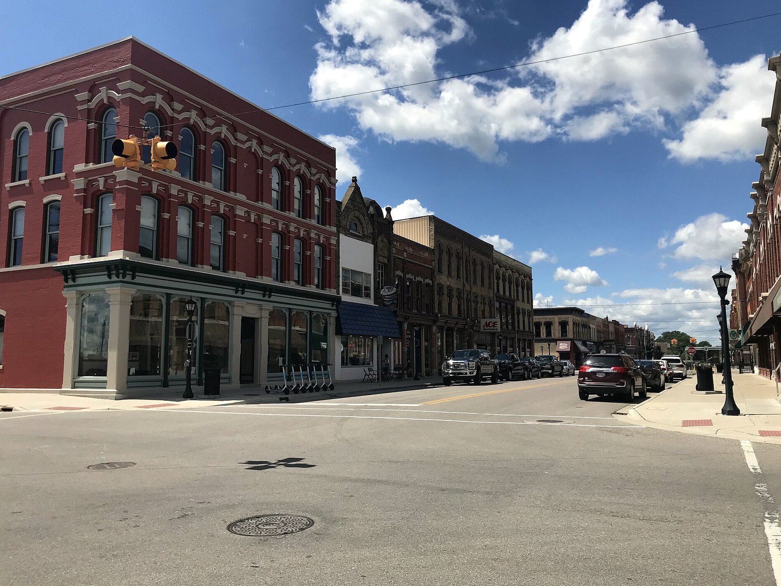A sunny corner on Midland Street in Bay City, with a three-story red brick building with arched windows and a green storefront, a row of older brick shops down the block, parked pickup trucks and a black lamppost under a blue sky with puffy clouds.