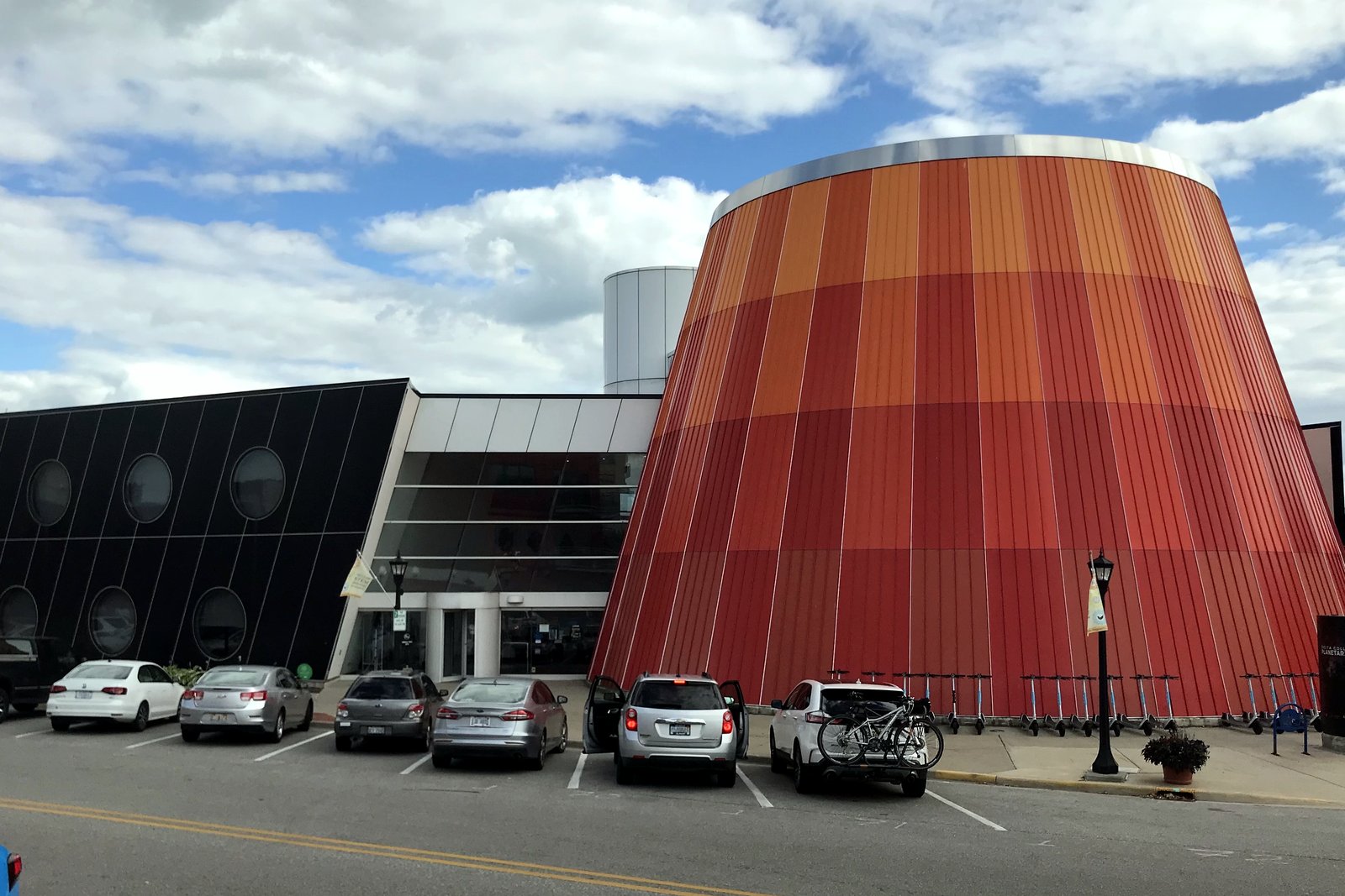 A tall, rounded building clad in vertical orange and red panels stands beside a lower dark building, with cars parked along the street in front under a partly cloudy sky.