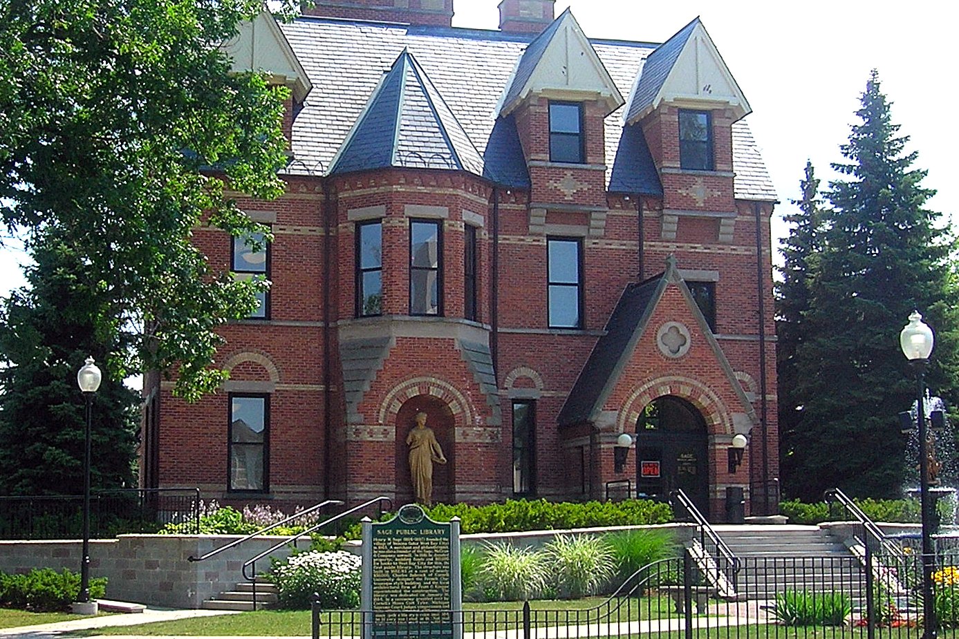 The Sage Branch Library in Bay City, a red brick Gothic Revival building with steep gray roofs, behind a black iron fence.