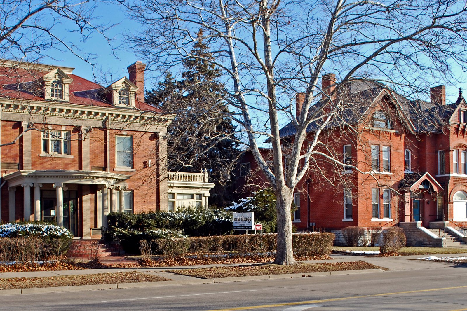 Two large 19th-century red brick houses with porches and tall windows sit behind bare trees on a residential avenue on a sunny winter day.