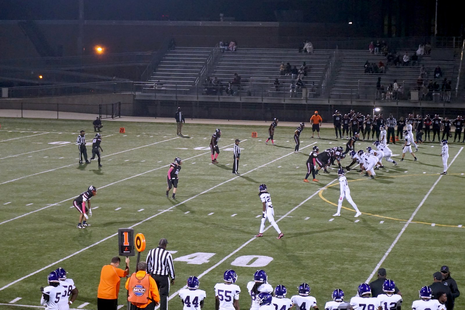 High school football players in black and white uniforms line up at night on lit artificial turf, with sideline players in the foreground and mostly empty bleachers behind.