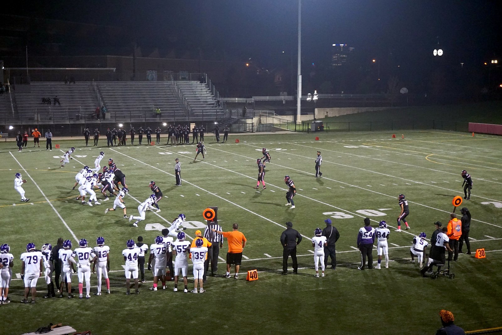 Football players line up at night on a lit artificial-turf field, with players on the sideline in the foreground and empty bleachers behind.