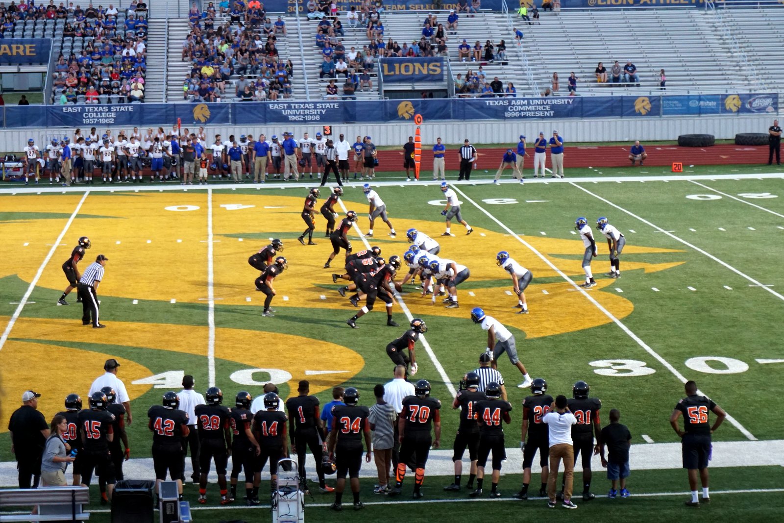High school football players in black jerseys and in white jerseys line up at the line of scrimmage on a gold-painted midfield logo as teammates watch from the sideline.