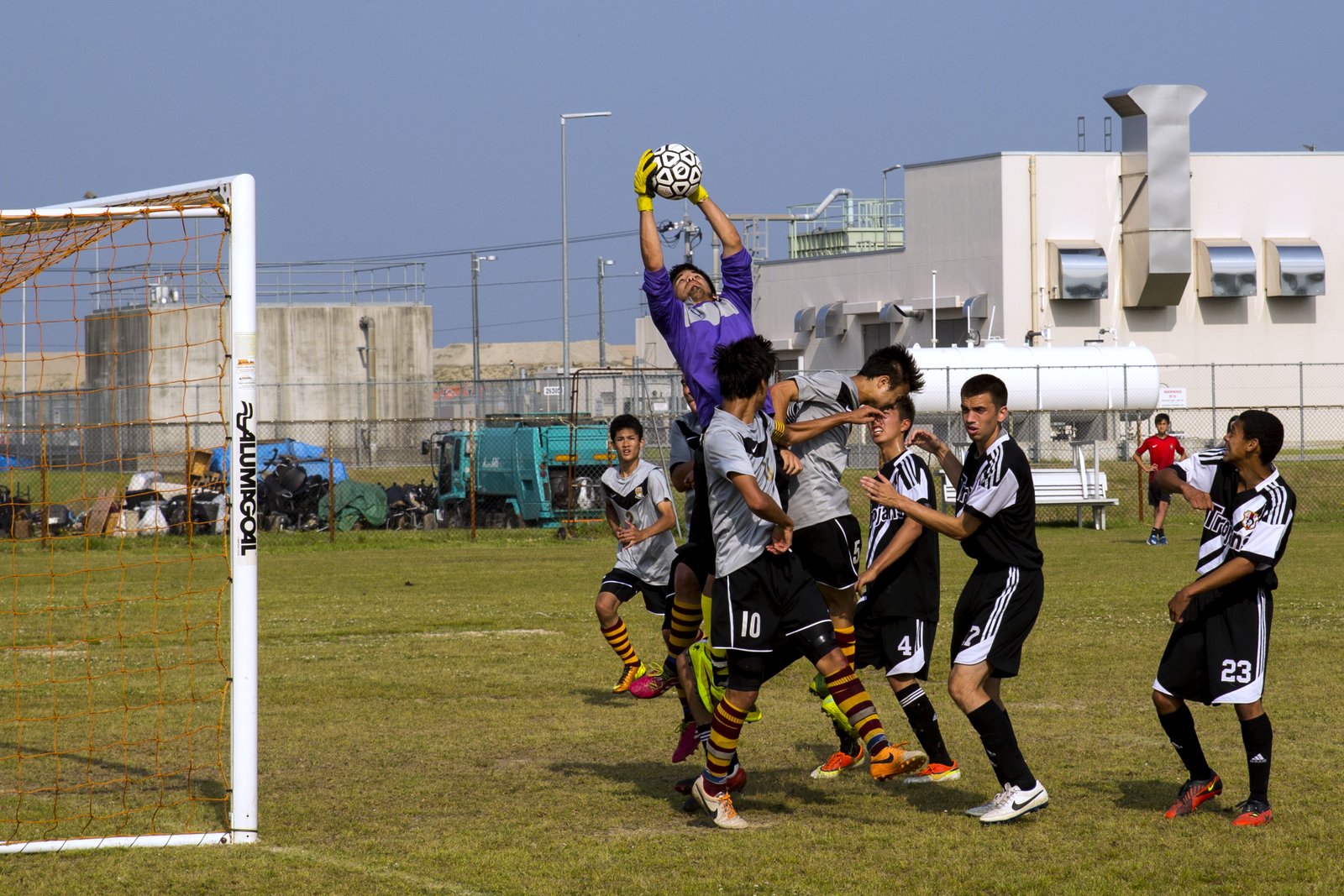 A high school goalkeeper in a purple jersey leaps above a crowd of players to catch a soccer ball in front of the net on a grass field.