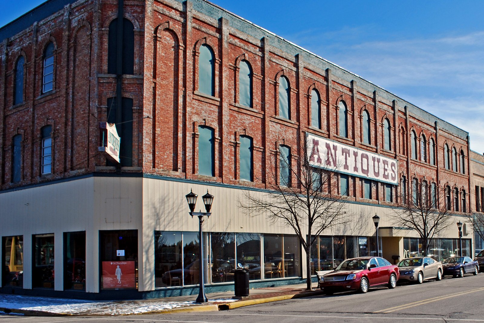 A three-story red-brick commercial building with tall arched windows and a large ANTIQUES sign at a corner in downtown Bay City, with cars parked along the street under a blue sky.