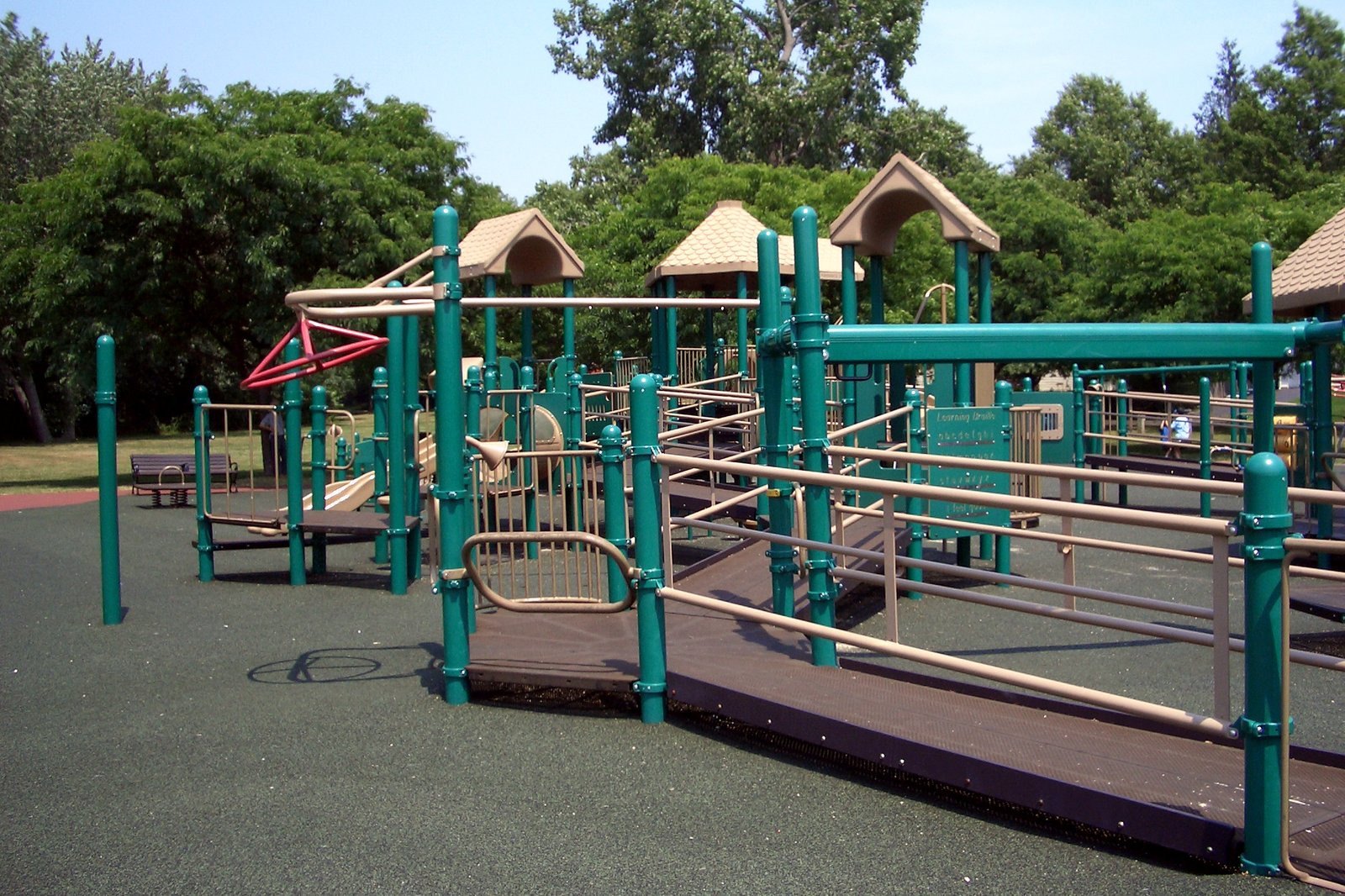 Green metal playground equipment with ramps, railings and tan peaked roofs stands on a rubber play surface in front of tall leafy trees.