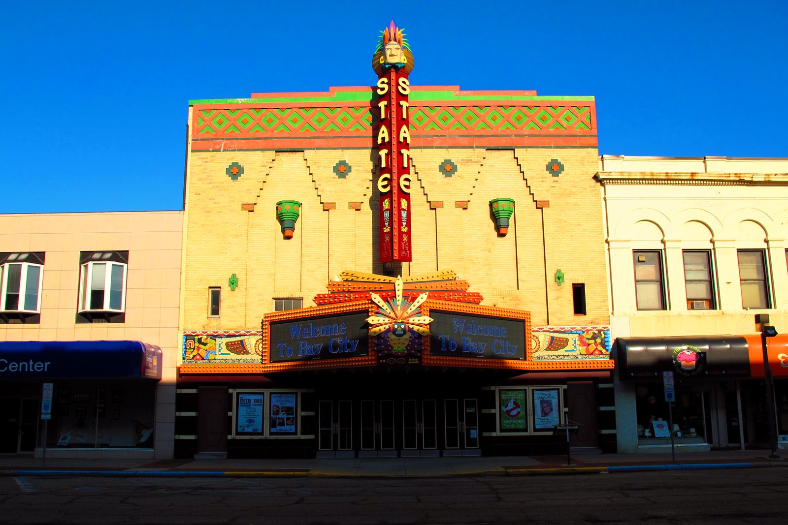 A theater with a tall vertical STATE sign and an ornate, colorful facade above its marquee, between two storefront buildings, under a blue sky.
