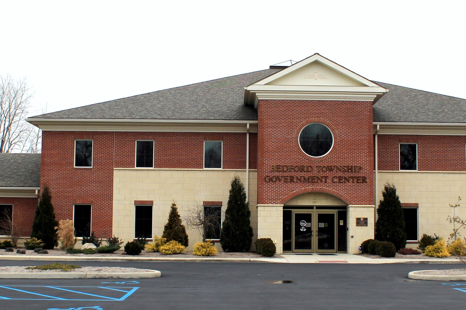 A two-story red-brick building with cream trim, a gabled center entrance with a round window, evergreen shrubs and a parking lot in front.
