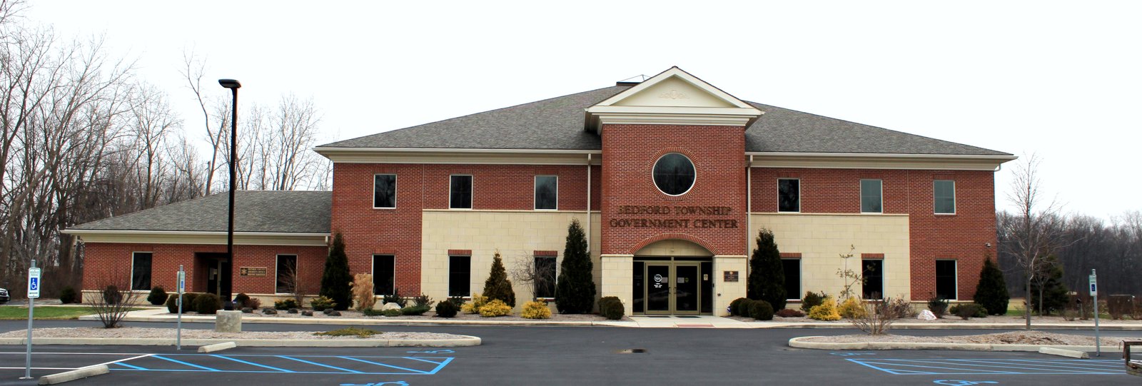 A two-story red-brick building with cream trim, a gabled center entrance with a round window, evergreen shrubs and a parking lot in front, with bare trees under an overcast sky.