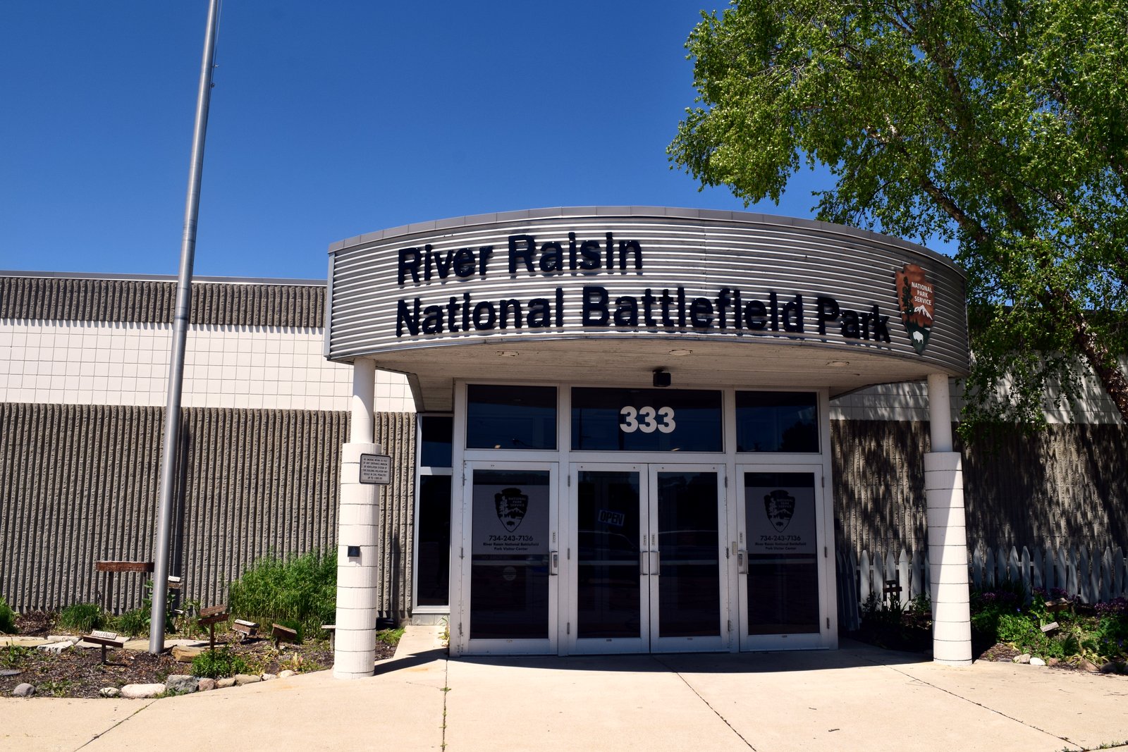 The entrance of a modern visitor center with a curved canopy reading River Raisin National Battlefield Park above glass doors marked 333, under a clear blue sky.