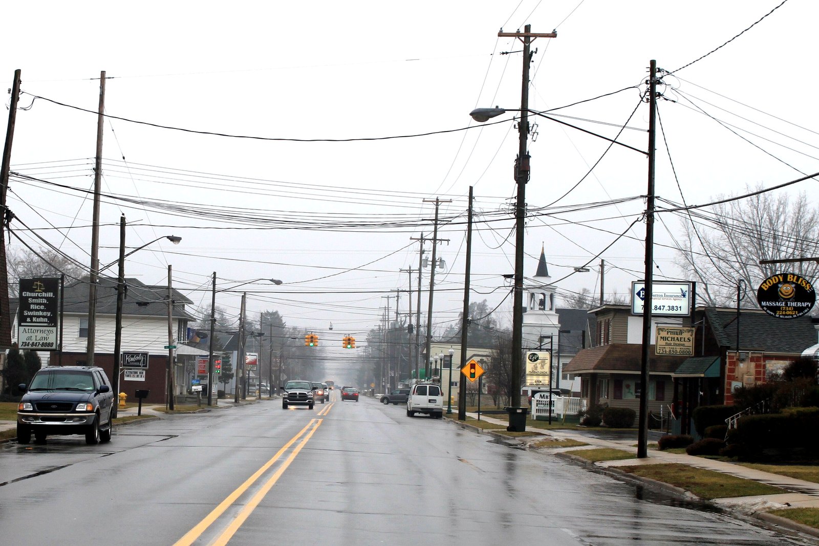 A two-lane commercial street lined with utility poles, small shops and parked cars runs south under an overcast sky.