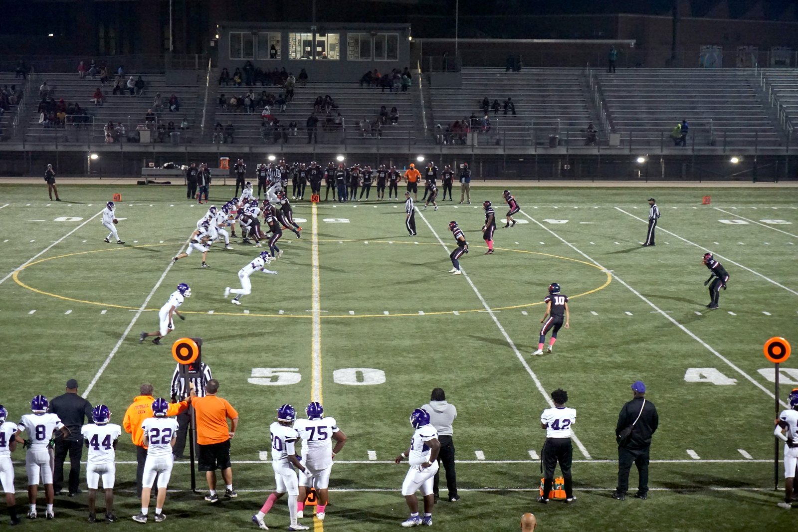 A high school football game at night under stadium lights, with players lined up on a green artificial-turf field and coaches and players watching from the sideline.
