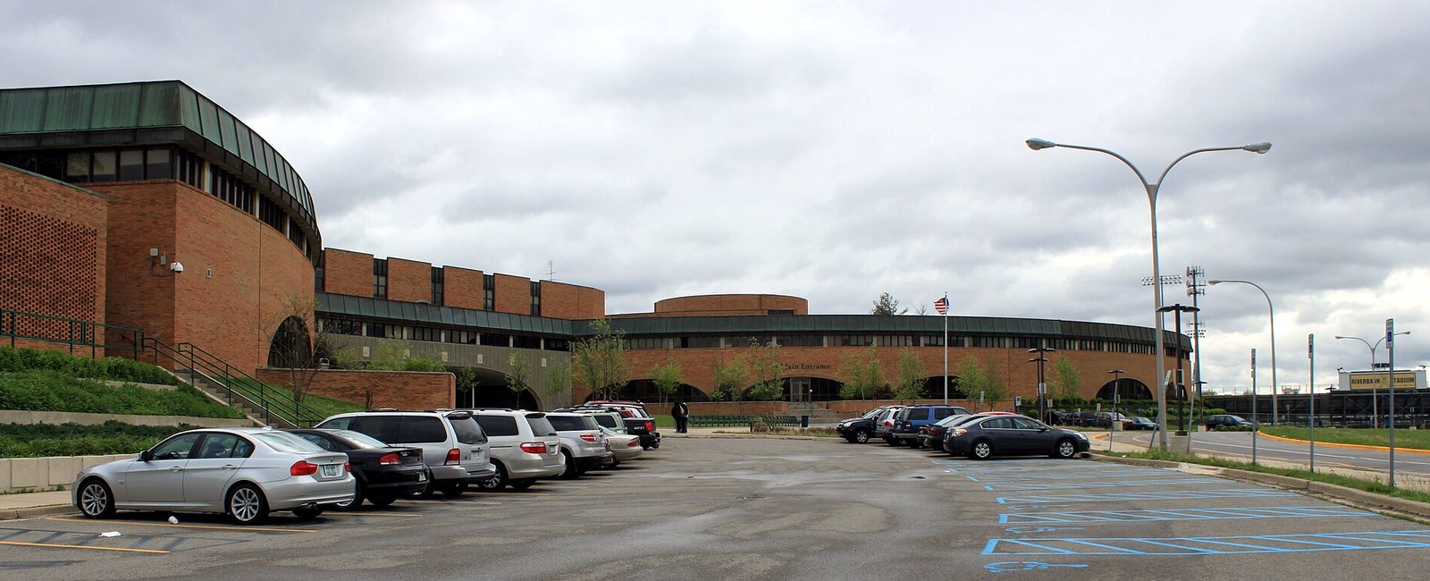 The long, low red-brick front of Huron High School in Ann Arbor, with curved green-roofed sections and a rounded central building, seen across a parking lot of parked cars under a heavy gray sky, with an American flag on a pole near the entrance.