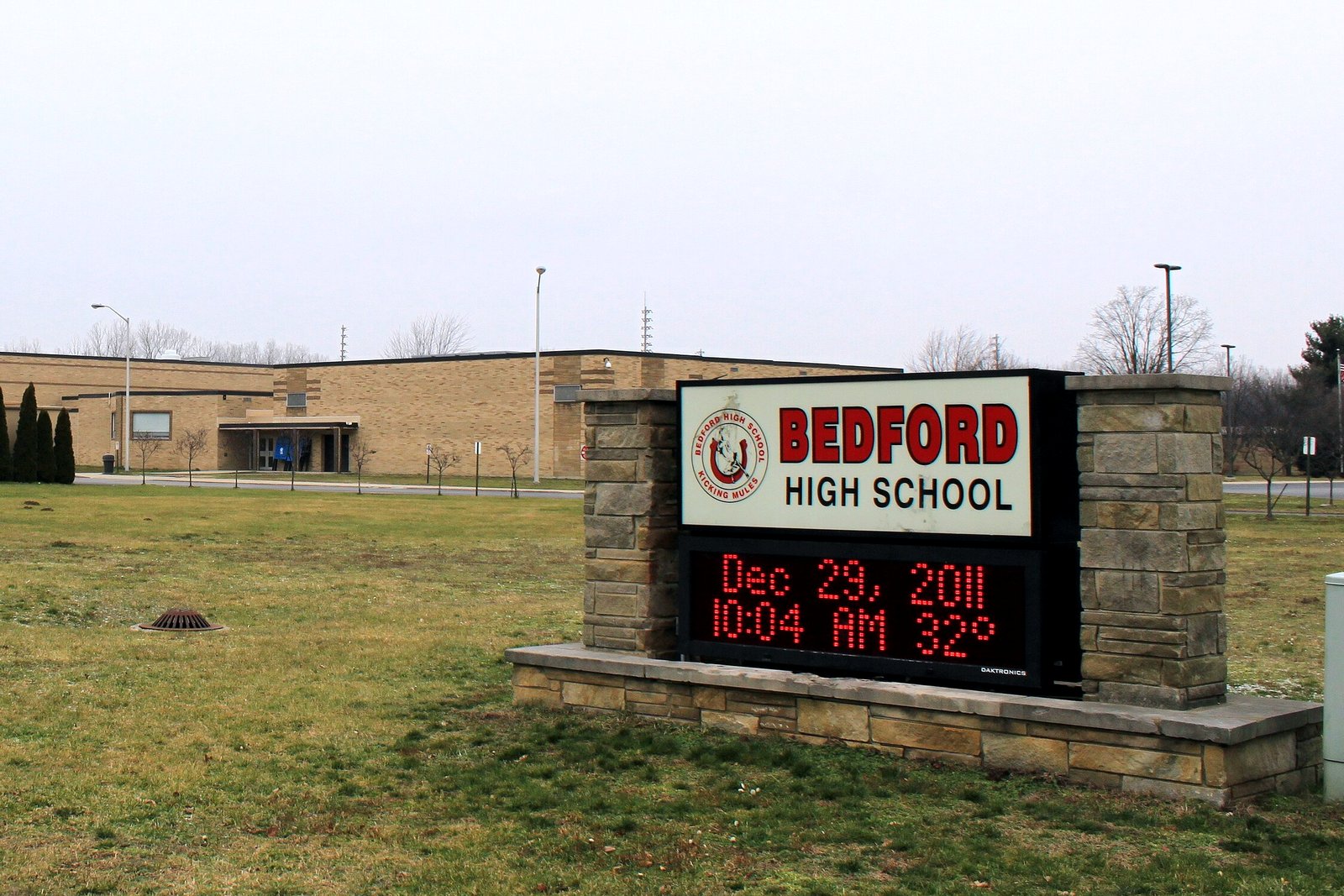 A long tan brick school building behind a wide lawn, with a stone-framed sign reading Bedford High School and a red electronic message board in the foreground.