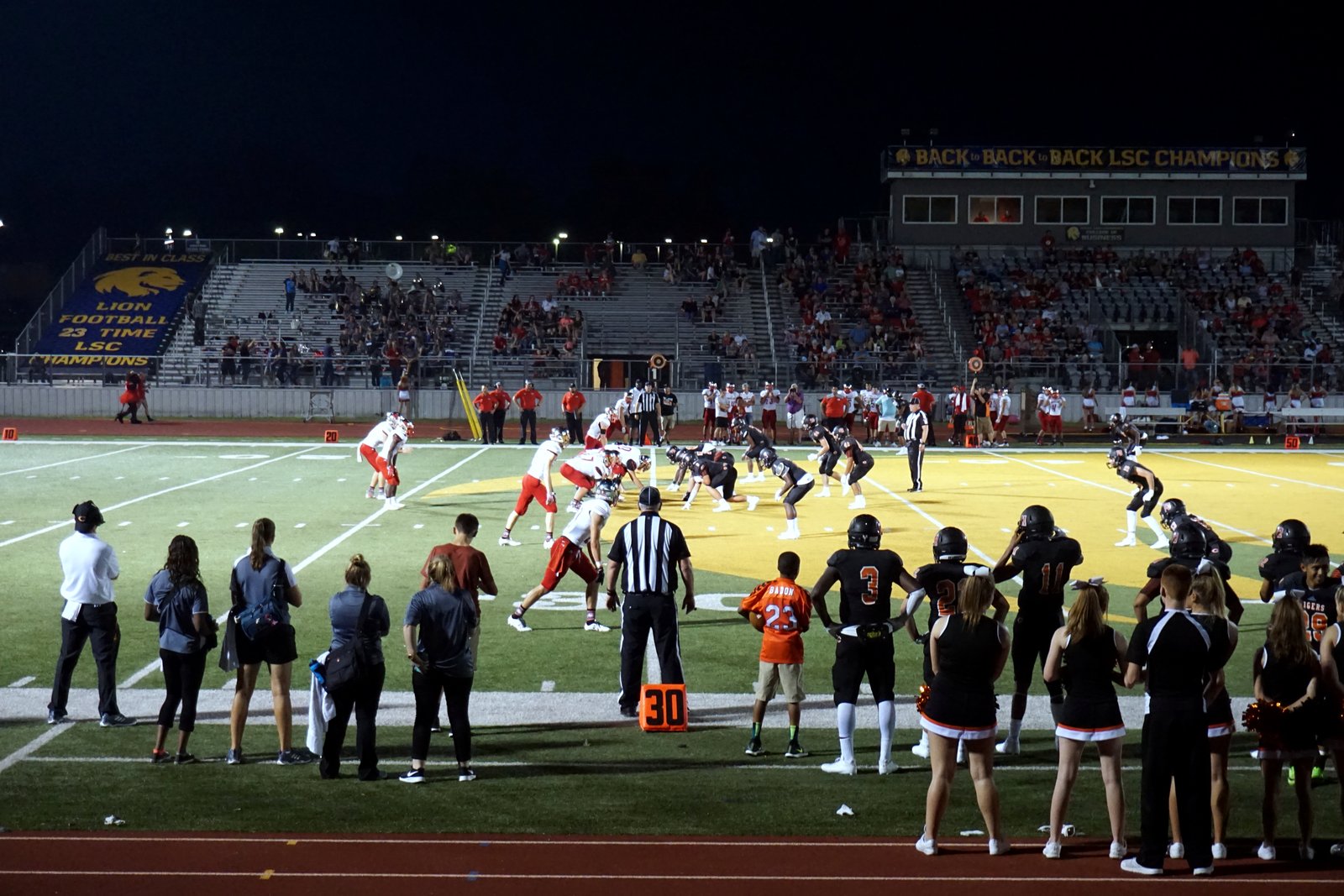 High school football players line up for a snap on a lighted field at night as cheerleaders, coaches and fans watch from the sideline and stands.