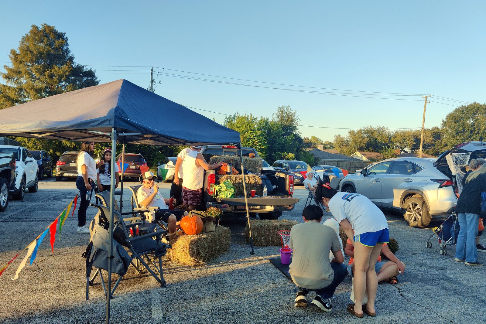A canopy tent, hay bales and a pumpkin beside a pickup truck in a parking lot, where adults and children gather around decorated car trunks under a clear evening sky.